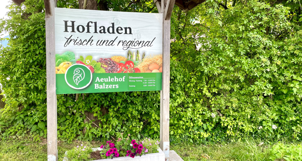 Schild des Hofladens Aeulehof mit Aufschrift „Frisch und regional“ vor grüner Hecke in Triesen.